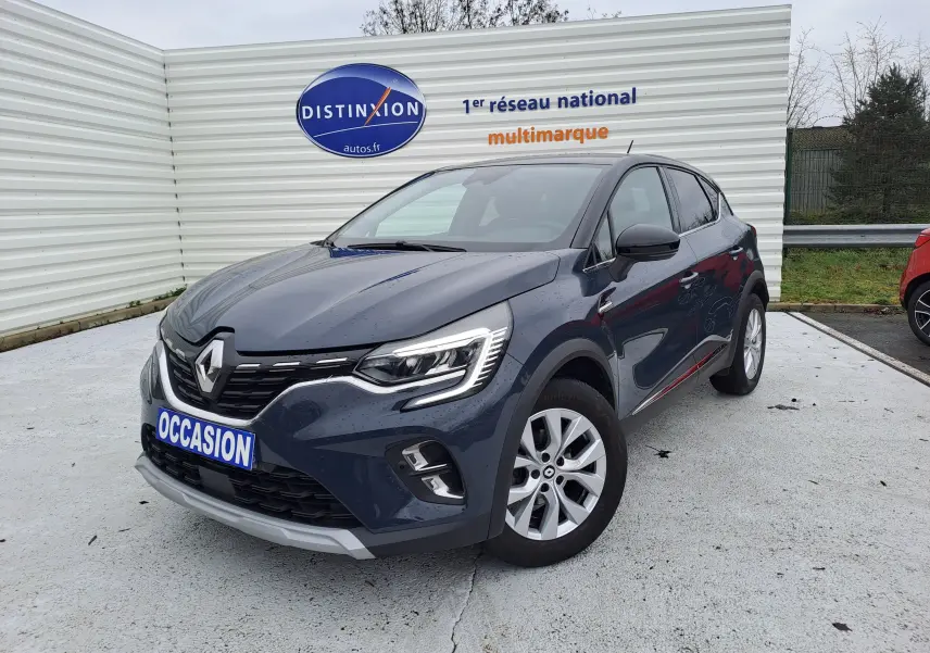 Renault Captur bleu marine en 3/4 avant droit, avec calandre distinctive et phares LED allumés, stationné devant un mur blanc.