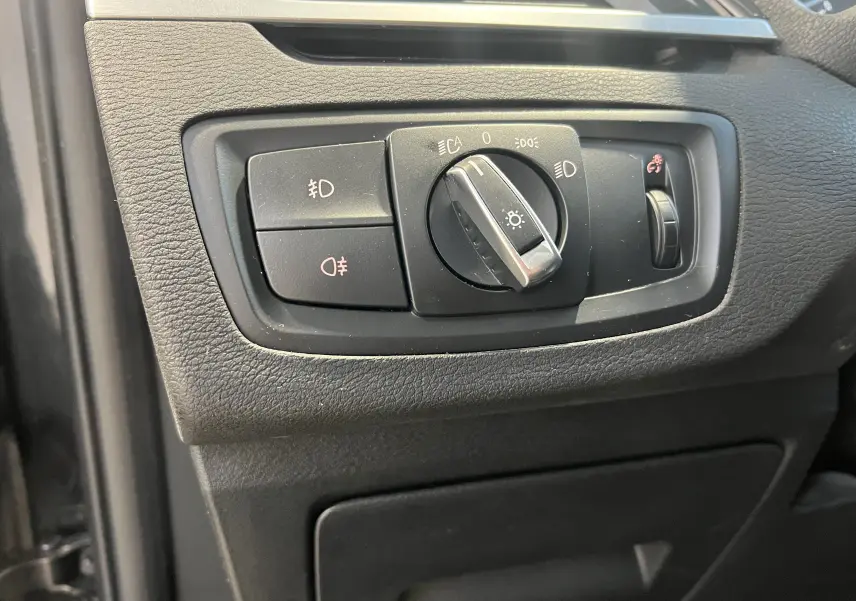 Gros plan sur la commande des phares noire du tableau de bord du BMW X1 F48 Saphirschwarz, côté gauche.