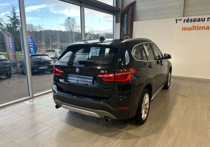 BMW X1 F48 noir saphir, vue 3/4 arrière droit en showroom avec logo X1 et double sortie d’échappement visible.