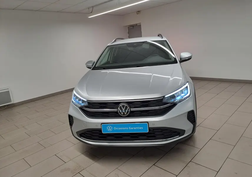 Vue avant d'un Volkswagen Taigo 2025 en peinture métallisée Reflet d'argent avec phares allumés dans un showroom.