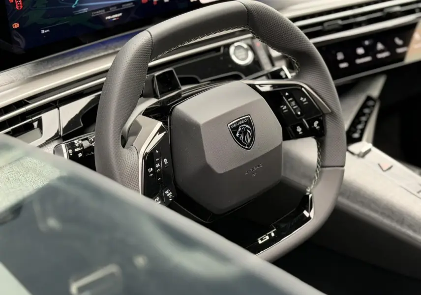 Gros plan sur le volant compact cuir perforé noir avec logo Peugeot et badge GT, tableau de bord numérique en arrière-plan.
