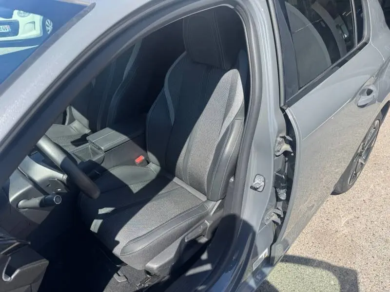 Vue intérieure côté conducteur d'une Peugeot 308 Hybrid gris 2025, siège réglable en hauteur et accoudoir central visibles.