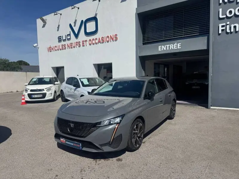 Peugeot 308 Hybrid gris vue 3/4 avant droit, jantes alliage 17 pouces, devant un garage avec enseigne sud VO.