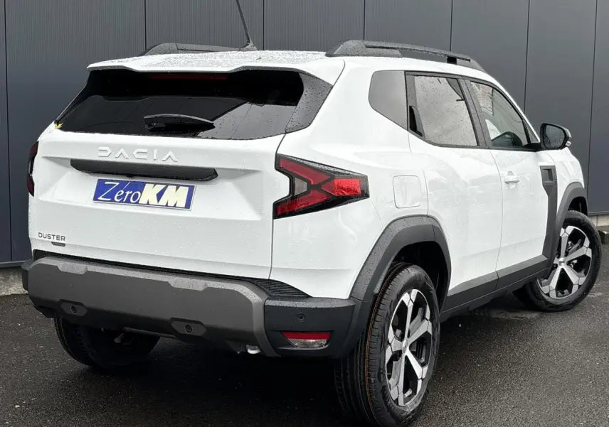 Vue 3/4 arrière droit du Dacia Duster blanc Glacier 2025 avec jantes alliage et barres de toit noires.