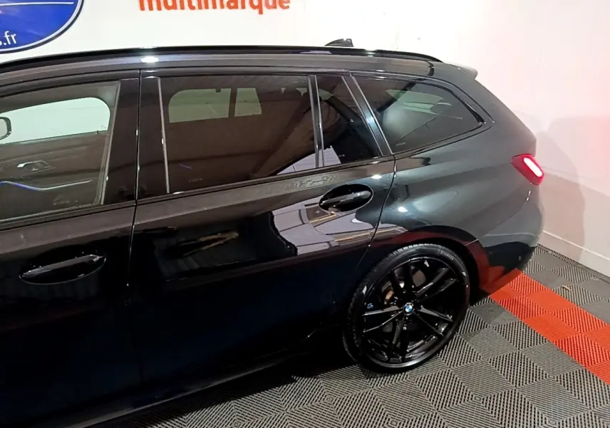 BMW Série 3 318i noire vue de profil côté gauche, avec jantes noires et feu arrière allumé.