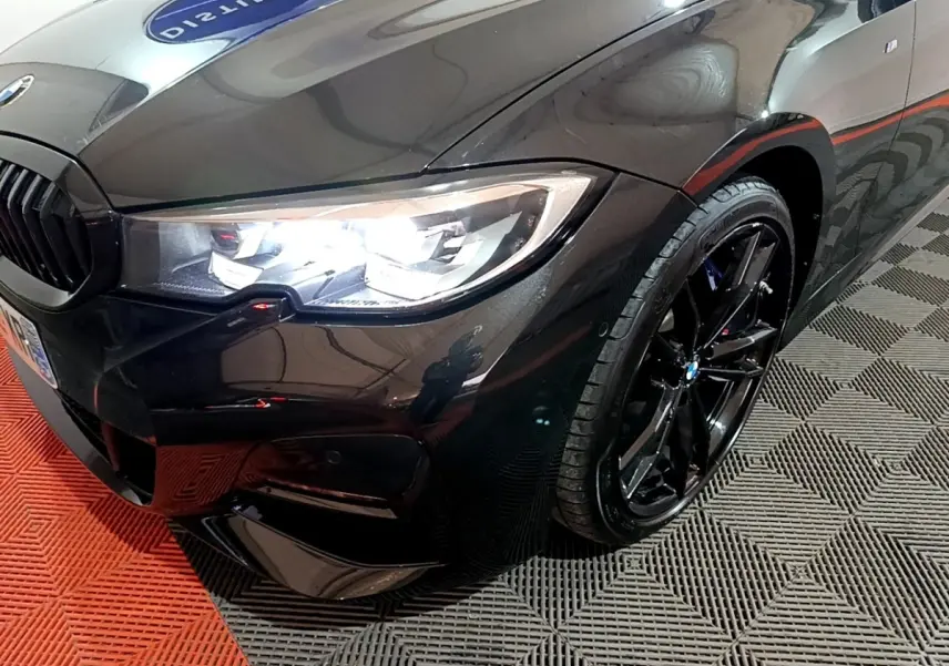 Vue 3/4 avant droit d'une BMW Série 3 318i noire avec jantes noires brillantes et phares LED allumés.