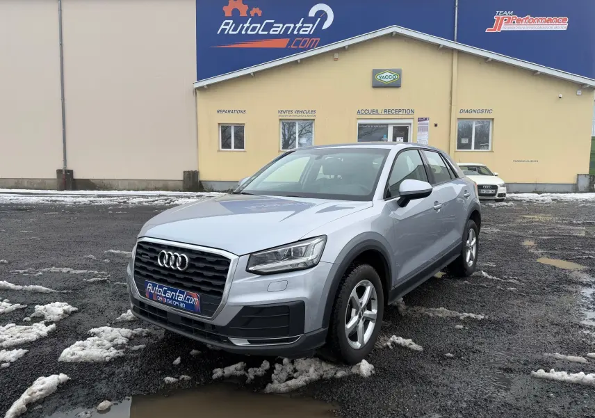 Audi Q2 gris aluminium vue 3/4 avant droit sur parking avec bâtiment AutoCantal en arrière-plan.