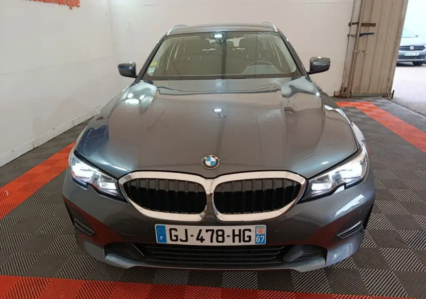 BMW Série 3 316d gris clair vue de face en intérieur, avec calandre double haricot et phares LED allumés.
