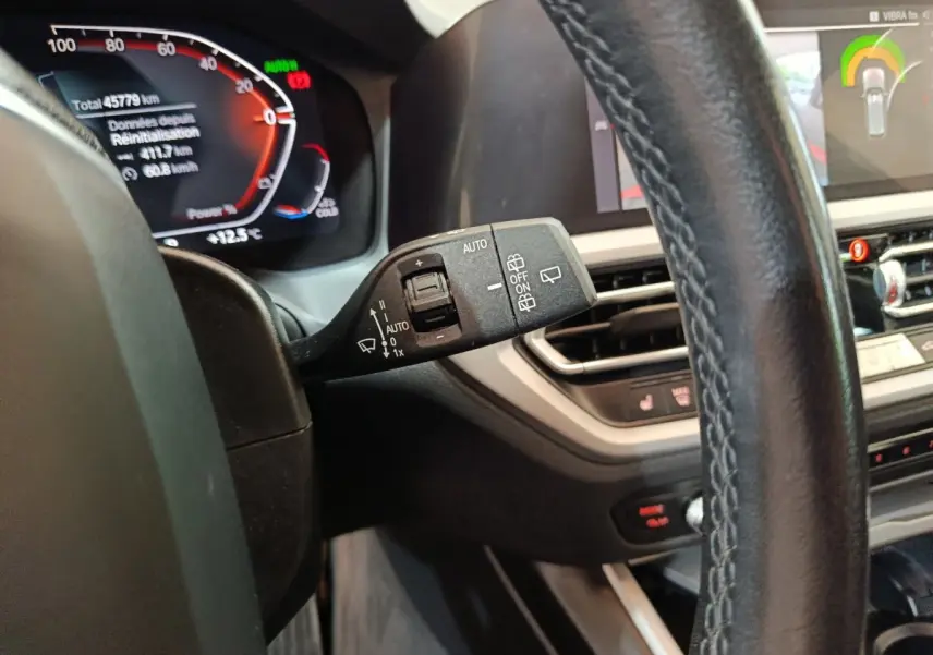 Gros plan sur le commodo de lève-glace à gauche du volant cuir d'une BMW Série 3 gris clair, tableau de bord numérique visible.