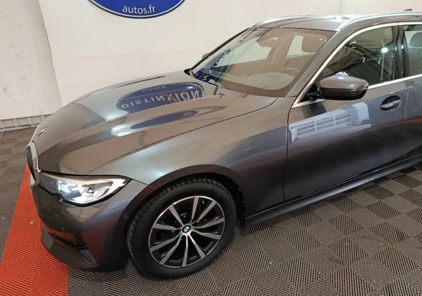 Vue 3/4 avant droit d'une BMW Série 3 gris clair avec phares LED allumés et jantes alliage noires.