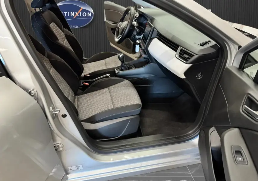 Vue intérieure côté conducteur de la Renault Clio V gris aluminium, montrant les sièges tissu bicolore et le tableau de bord moderne.