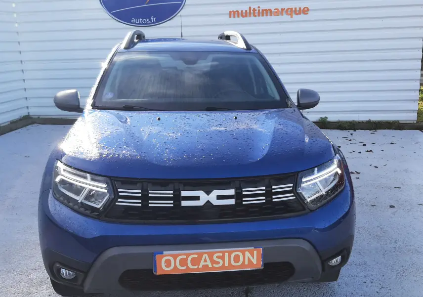Vue avant d'un Dacia Duster bleu 2023 avec calandre noire et phares LED, plaque "OCCASION" visible.