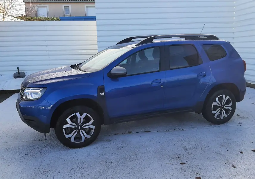 Vue de profil côté gauche d'un Dacia Duster bleu 2023 avec barres de toit et jantes bi-ton noires et argentées.