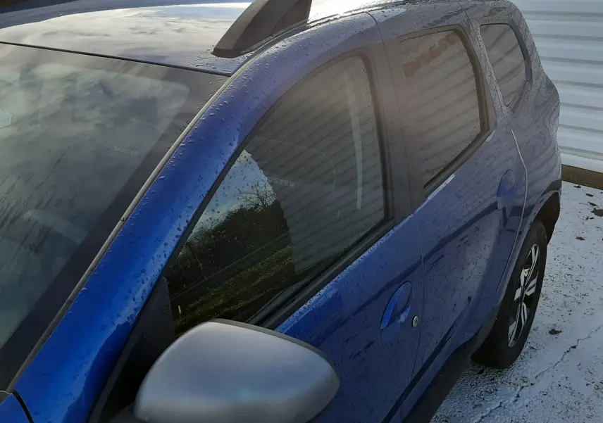 Vue 3/4 arrière droite d'un Dacia Duster bleu 2023 avec barres de toit noires et rétroviseur gris.