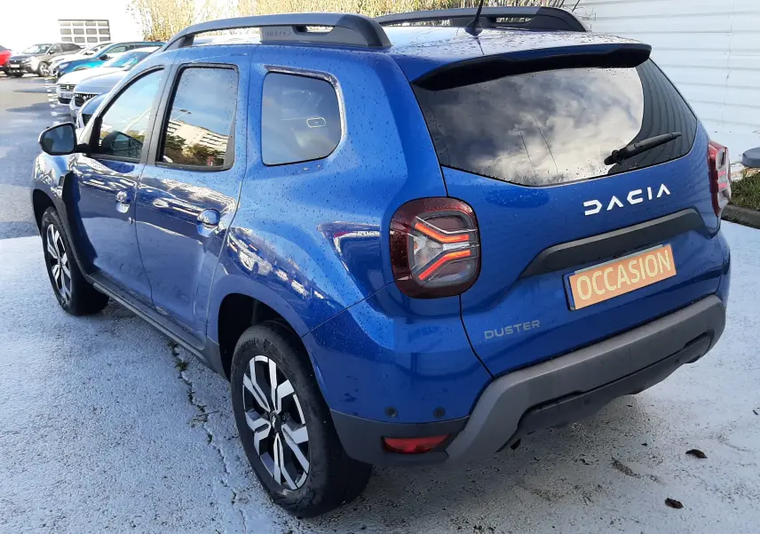 Vue 3/4 arrière droite d'un Dacia Duster bleu 2023 avec toit noir, barres de toit et feux arrière LED distinctifs.