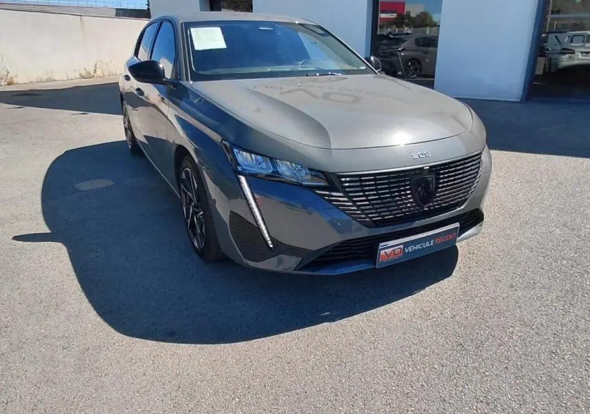 Peugeot 308 gris vue 3/4 avant droit, avec calandre noire distinctive et feux LED verticaux allumés.