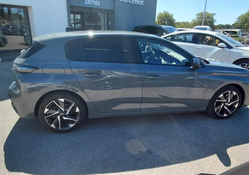 Peugeot 308 gris vue de profil côté gauche, stationnée en extérieur devant un bâtiment blanc.