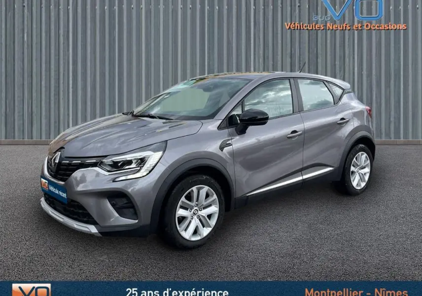 Vue 3/4 avant droit d'un Renault Captur gris 2021 avec jantes alliage et rétroviseurs noirs sur fond industriel.