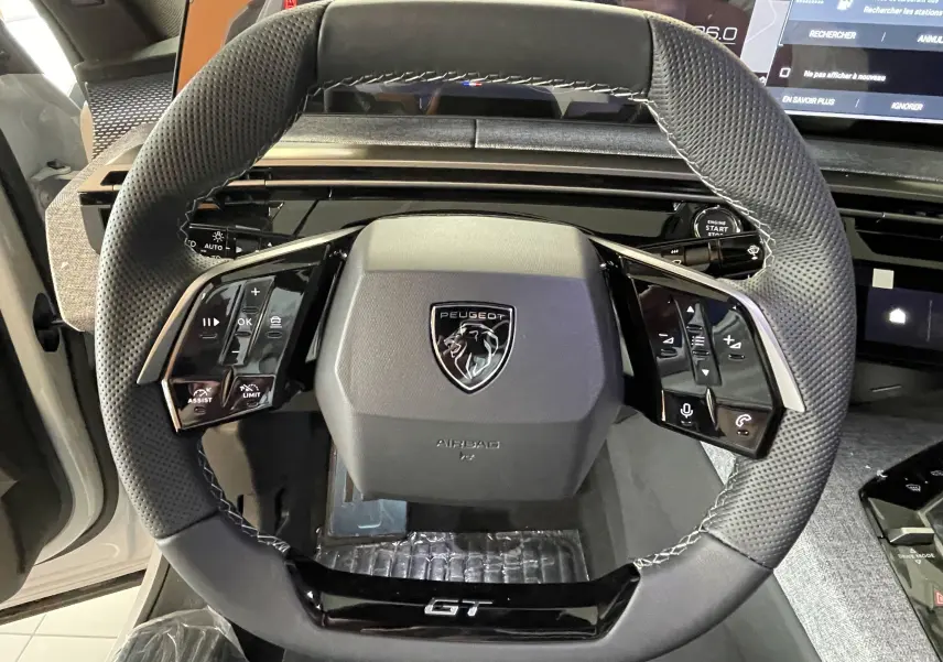 Vue rapprochée du volant GT noir perforé avec logo Peugeot au centre dans l'habitacle du 3008 Hybrid blanc.