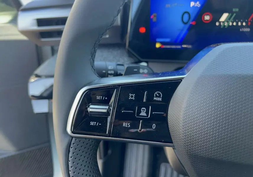 Gros plan sur les commandes au volant à gauche de la Renault Clio blanc glacier, avec affichage numérique en arrière-plan.