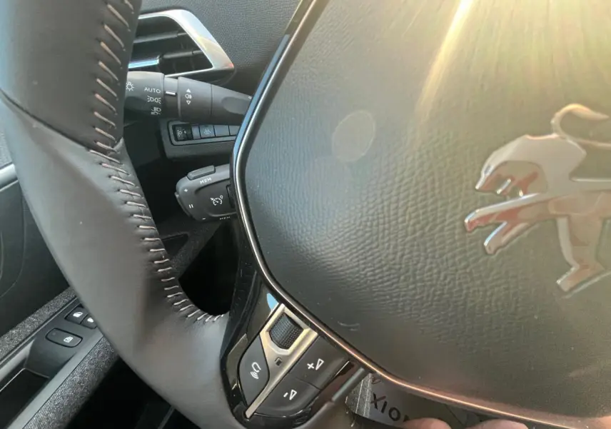 Gros plan sur le volant cuir noir du Peugeot 3008 gris Artense avec commandes intégrées, lumière naturelle entrant côté gauche.