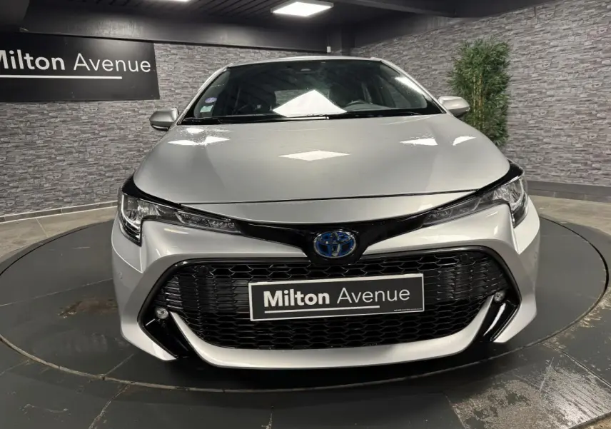Vue frontale d'une Toyota Corolla hybride gris métallisé avec calandre noire et phares LED dans un showroom.