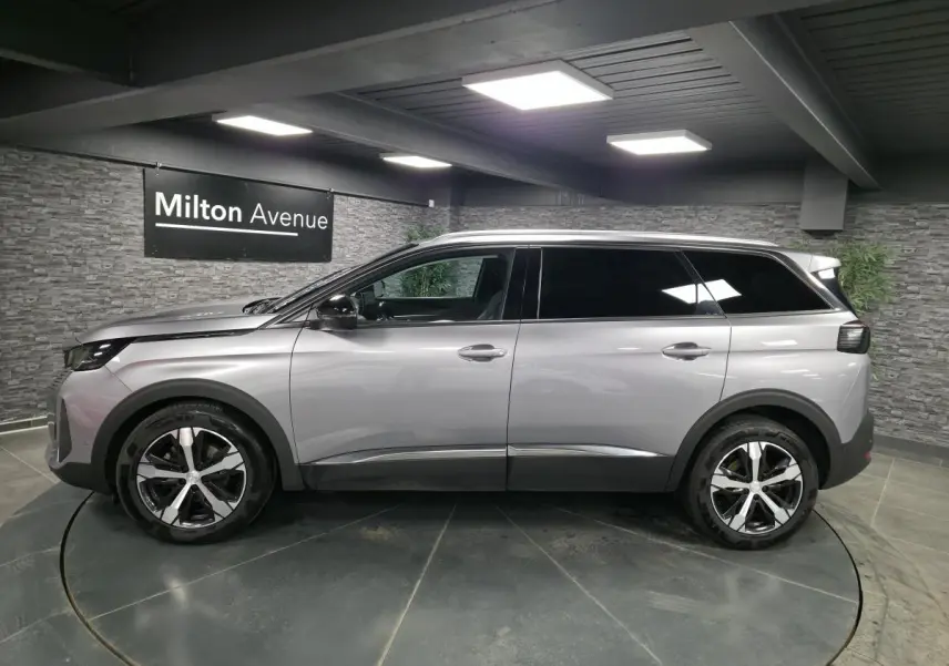 Profil côté gauche du Peugeot 5008 gris métallisé 2022 avec jantes alliage bi-ton et vitres surteintées dans un showroom.