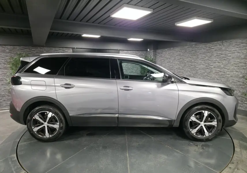 Profil côté gauche d'un Peugeot 5008 gris métallisé avec jantes alliage bi-ton et vitres surteintées en intérieur showroom.