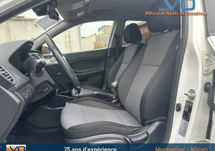 Vue intérieure côté conducteur de la Hyundai i20 blanche 2019, sièges tissu gris et volant réglable visible.