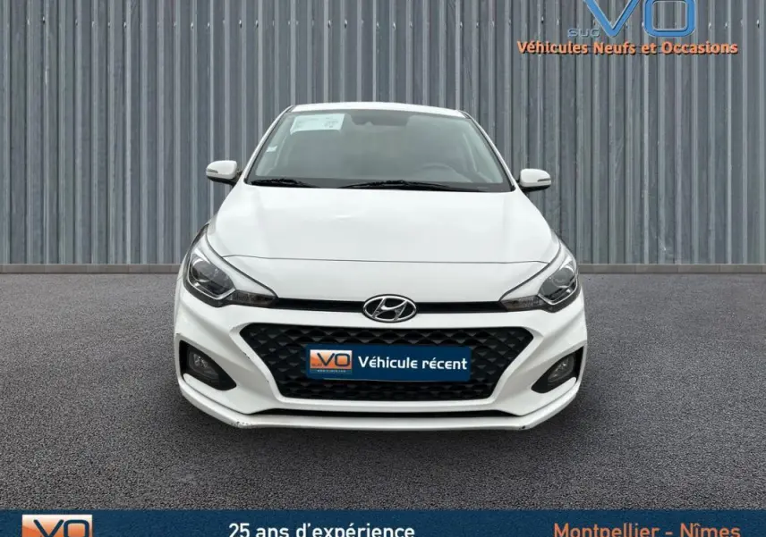 Vue frontale d'une Hyundai i20 blanche de 2019 avec calandre noire et phares LED allumés.