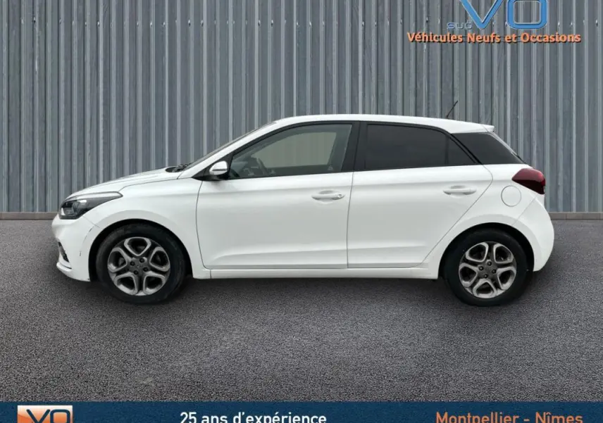 Profil côté gauche d'une Hyundai i20 blanche de 2019 avec jantes alliage et vitres teintées sur fond industriel.