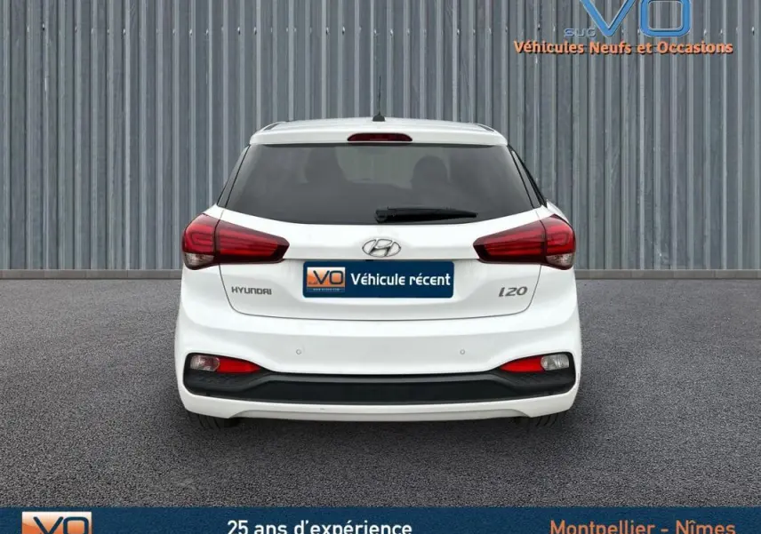 Vue arrière d'une Hyundai i20 blanche de 2019 avec feux rouges et lunette arrière teintée sur fond industriel.