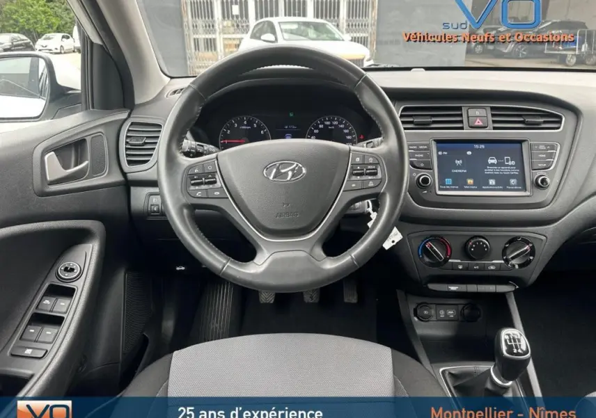 Vue intérieure frontale du tableau de bord et volant d'une Hyundai i20 blanche 2019 avec boîte manuelle et écran tactile central.