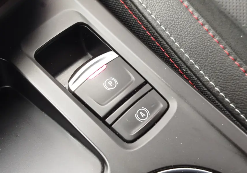 Gros plan sur les boutons de frein à main électronique et arrêt automatique du moteur, avec surpiqûres rouges sur cuir noir.