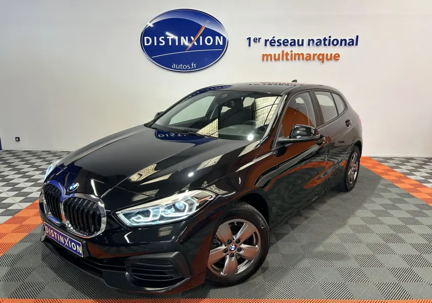 BMW Série 1 16d noire vue en 3/4 avant droit, phares LED allumés et jantes alliage visibles dans un showroom.