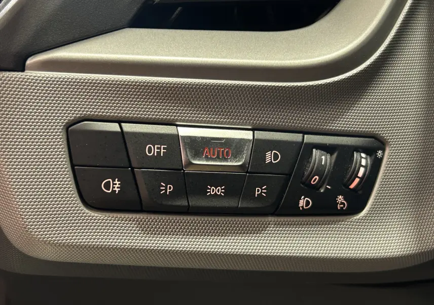Gros plan sur les commandes d'éclairage de la BMW Série 1 16d 2023, avec bouton AUTO et réglages lumineux sur fond texturé gris.
