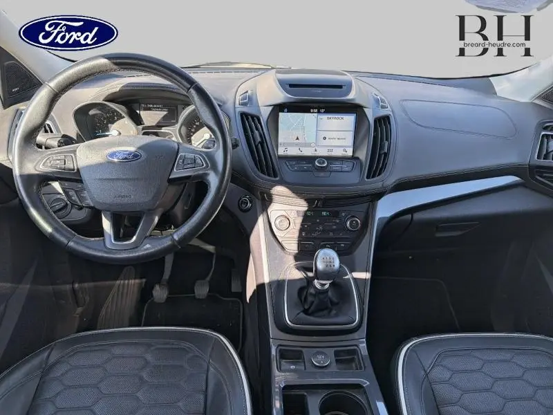 Vue intérieure avant du Ford Kuga 2017 avec tableau de bord noir, écran GPS central et volant cuir multifonction.