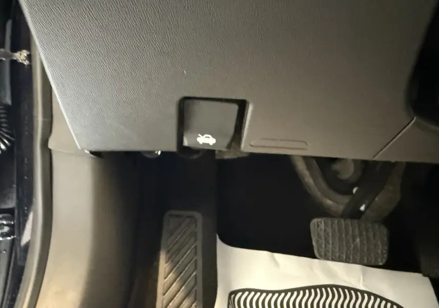 Le levier d'ouverture du capot situé sous le tableau de bord côté gauche, avec les pédales de frein et d'accélérateur visibles.