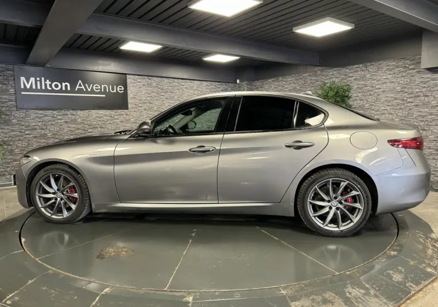 Profil droit d'une Alfa Romeo Giulia gris métallisé avec jantes alliage 18 pouces et vitres arrière surteintées dans un showroom.