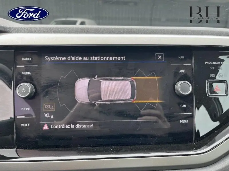 Écran tactile montrant l'aide au stationnement d'une Volkswagen Polo blanche vue de dessus, avec alertes de distance.