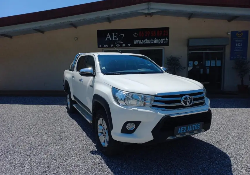 Toyota Hilux 4x4 blanc en 3/4 avant droit, double cabine, avec calandre chromée et barres de toit visibles.