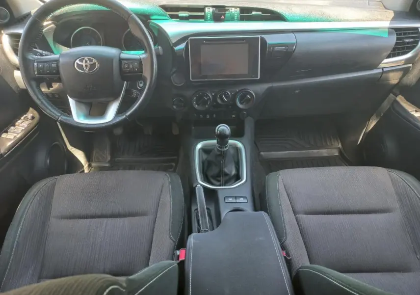 Vue intérieure avant du Toyota Hilux 2018, tableau de bord noir avec volant multifonction et levier de vitesse manuel.