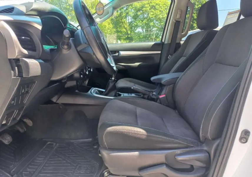 Vue intérieure côté conducteur du Toyota Hilux blanc 2018, montrant les sièges noirs et la boîte manuelle.