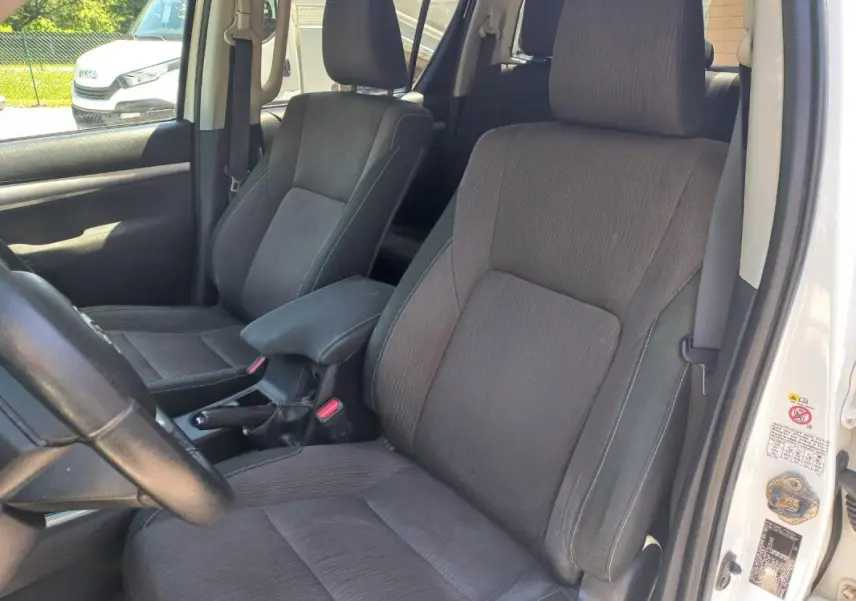 Vue intérieure côté conducteur du Toyota Hilux blanc 2018, montrant les sièges en tissu gris et la boîte manuelle.
