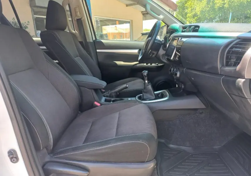 Vue intérieure côté conducteur du Toyota Hilux blanc 2018, mettant en valeur les sièges noirs et la boîte manuelle.