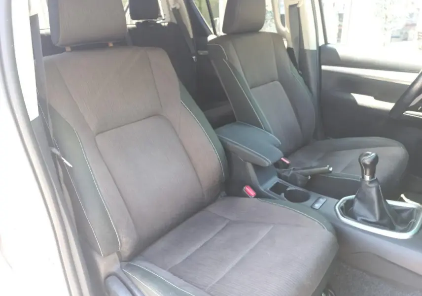 Vue intérieure côté conducteur du Toyota Hilux blanc 2018, montrant les sièges en tissu gris et la boîte manuelle.