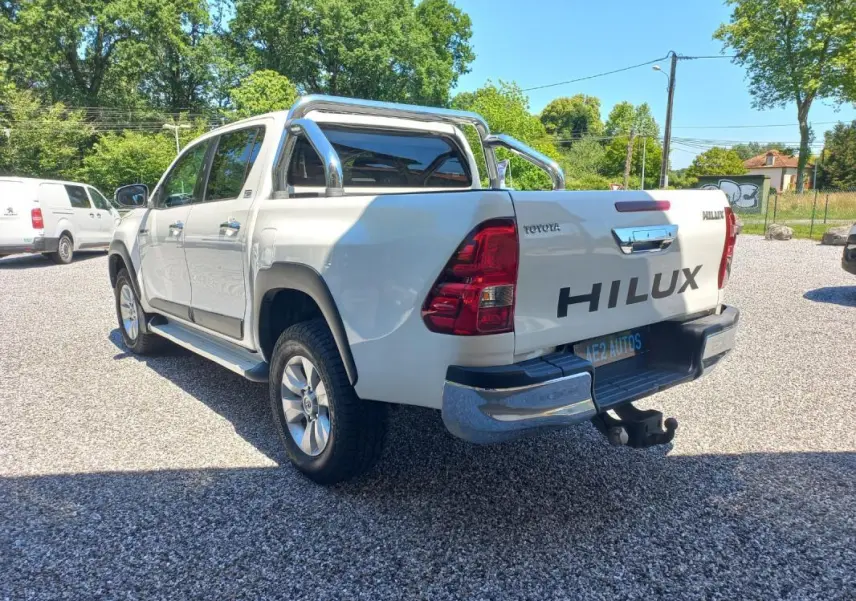 Toyota Hilux 2018 blanc en 3/4 arrière droit, double cabine avec barres chromées et porte-remorque visible.