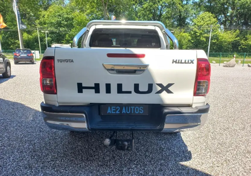 Vue arrière d'un Toyota Hilux blanc 4x4 avec hayon marqué HILUX et attelage visible, stationné en extérieur.