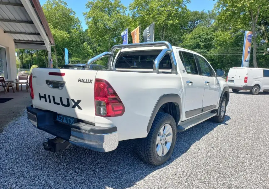Toyota Hilux blanc en 3/4 arrière droit, double cabine avec barre de protection chromée sur benne, pneus tout-terrain visibles.