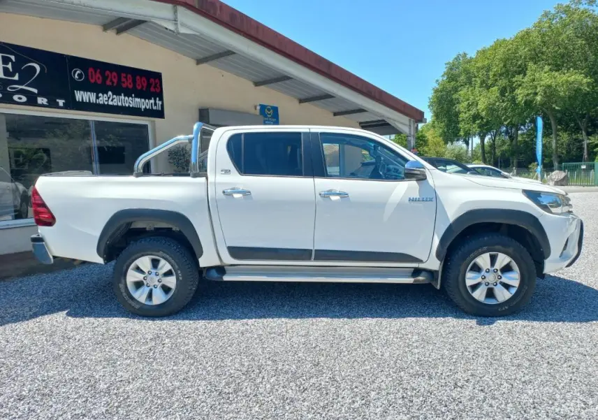 Profil côté gauche du Toyota Hilux 4x4 blanc double cabine avec barre de protection chromée, roues tout-terrain visibles.